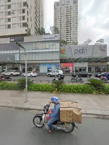 A street scene in Thao Dien, Saigon, featuring a motorcyclist with a delivery box, multi-story buildings, a pedestrian walkway, and vehicles. The back