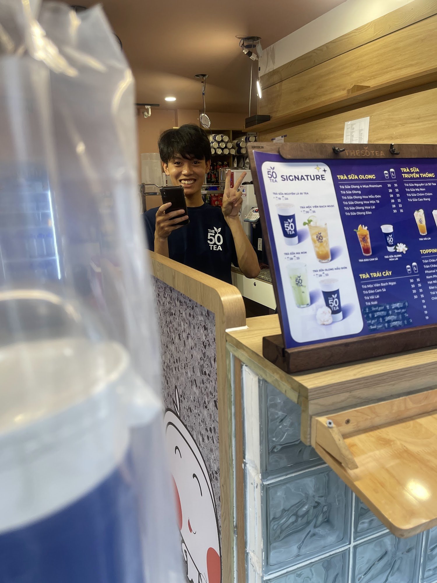 A person is taking a selfie in front of the menu board at 'The 50 Tea' store located inside Thao Dien, Saigon.