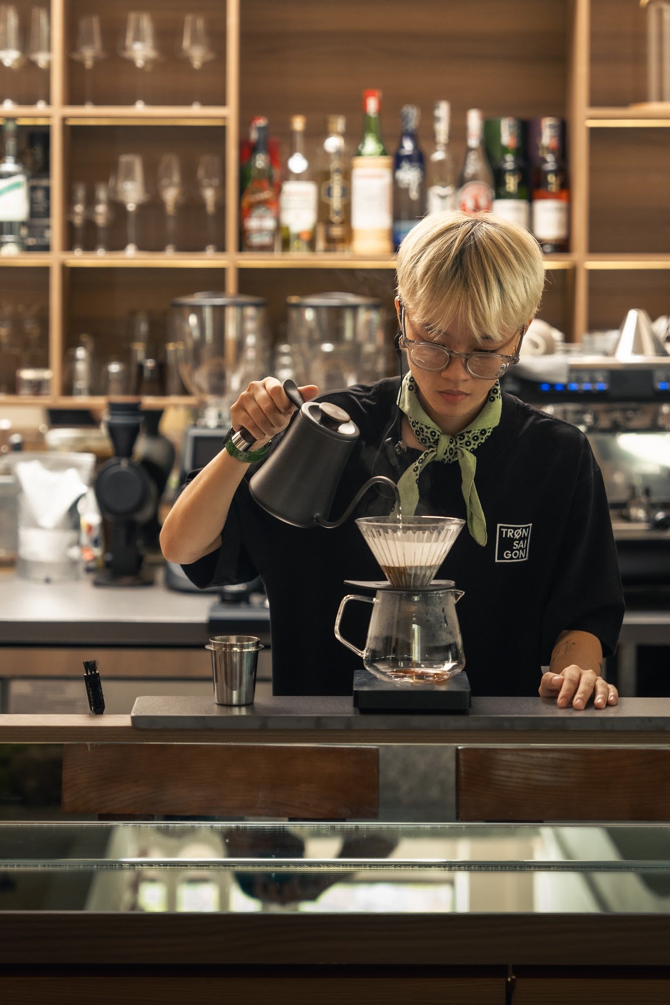 A barista is pouring hot water into a Chemex coffee dripper on the counter of an upscale bar or café, with various bottles and glasses in the backgrou