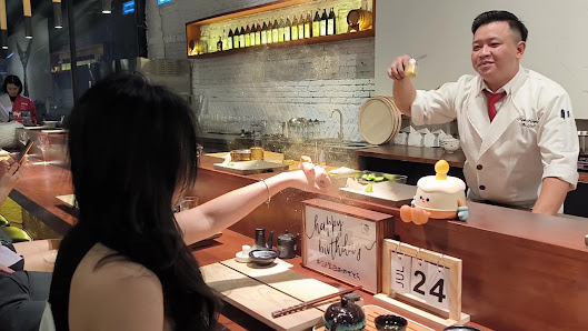 A restaurant interior in Thao Dien, Saigon. A chef in a white uniform with a red tie is interacting with a customer. The counter features a 'Happy Bir