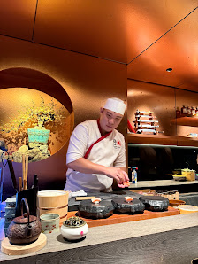 A chef in a white uniform with a red apron and white hat is preparing food at a counter. The counter has various items like small bowls, a teapot, and