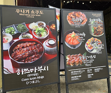 The image shows two large menu boards displaying various Korean dishes such as Napa cabbage kimchi and Com lron nhit, with prices listed in Vietnamese