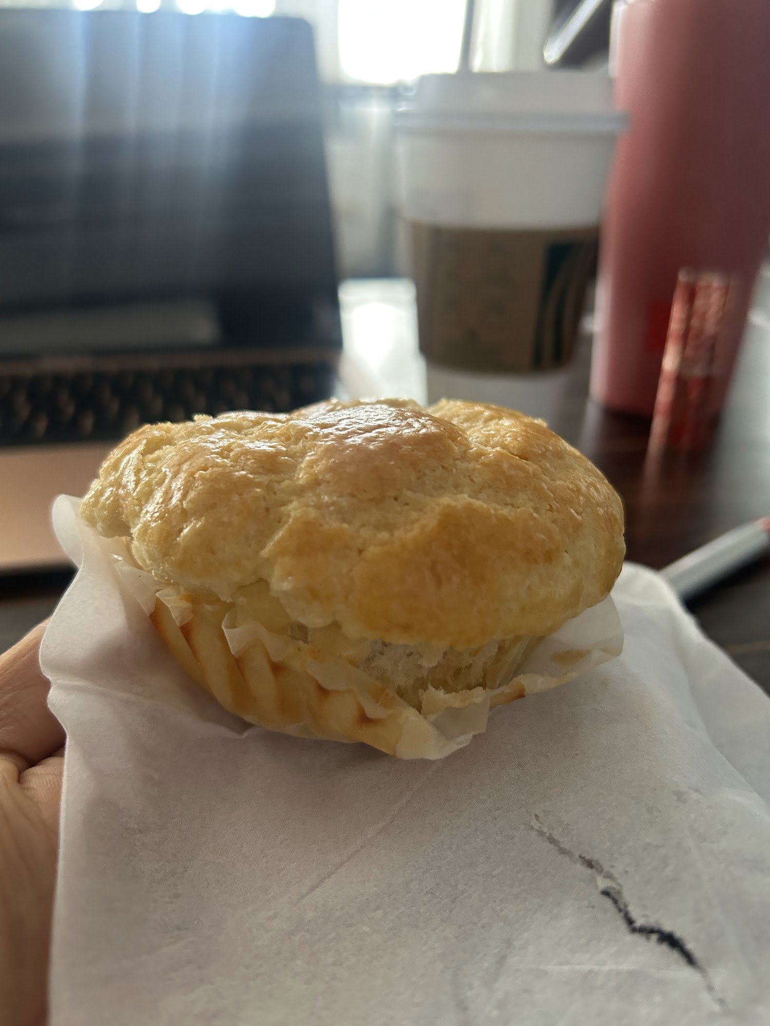 A close-up of a golden-brown pastry on parchment paper held in someone's hand with various items blurred out in the background, including what appears