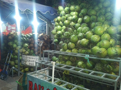 The image shows a market stall at night selling green coconuts under bright lights.