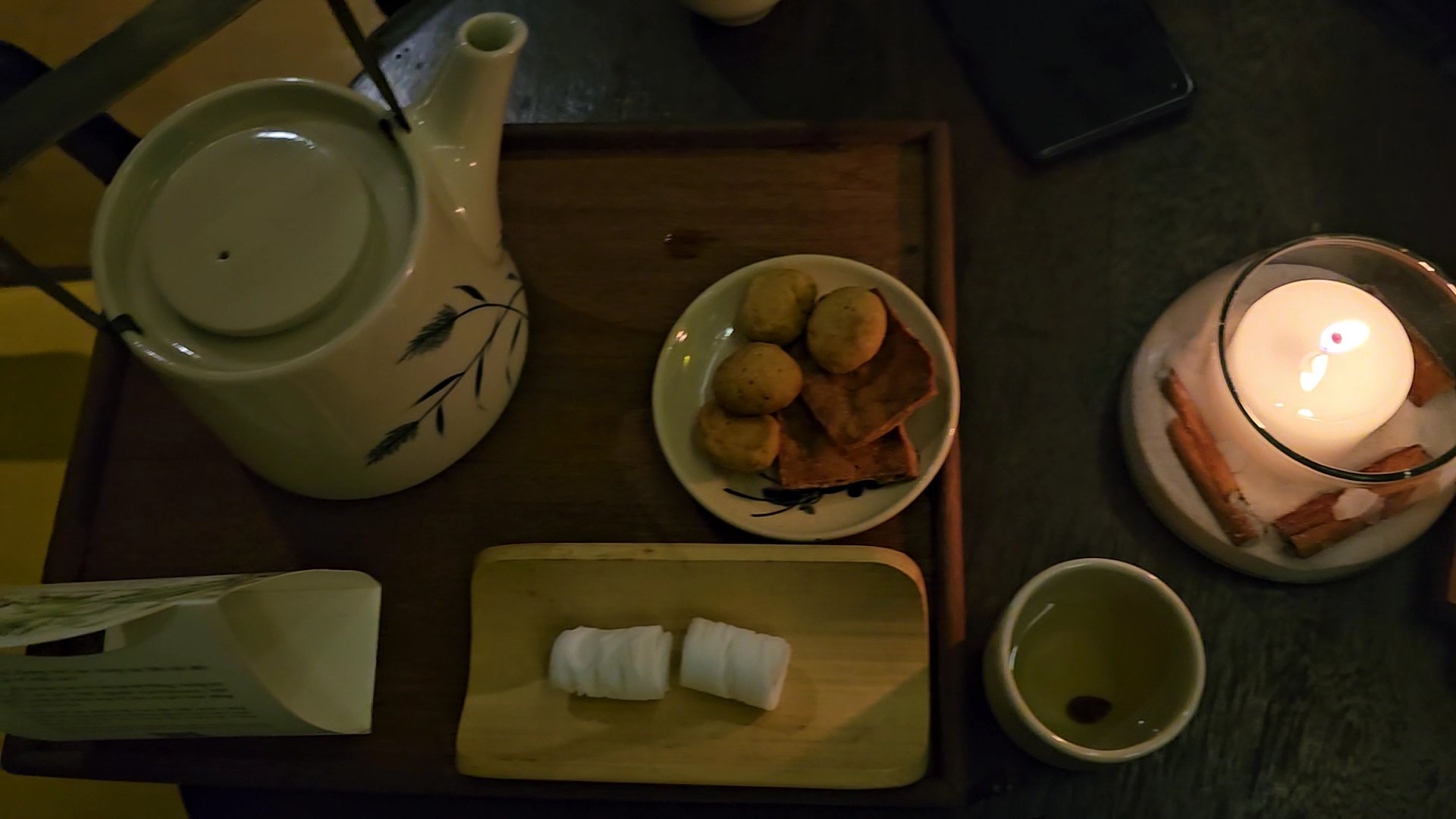 The image shows a dimly lit interior setting featuring a wooden tray on which various items are placed for what appears to be a tea or coffee break, i