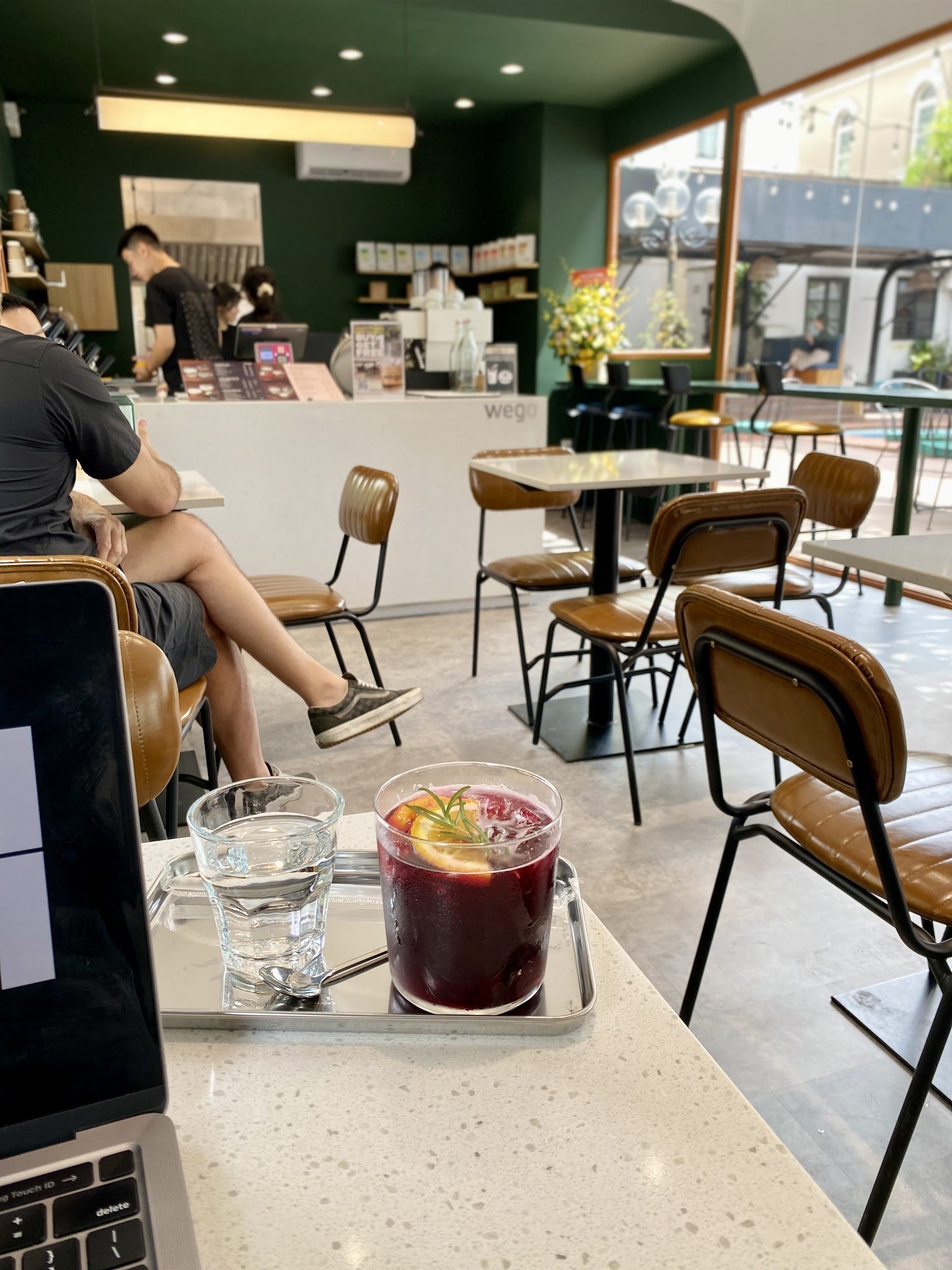 The image shows an interior view of a modern café or coffee shop. A person is seated at a table with a laptop and two glasses, one containing water an