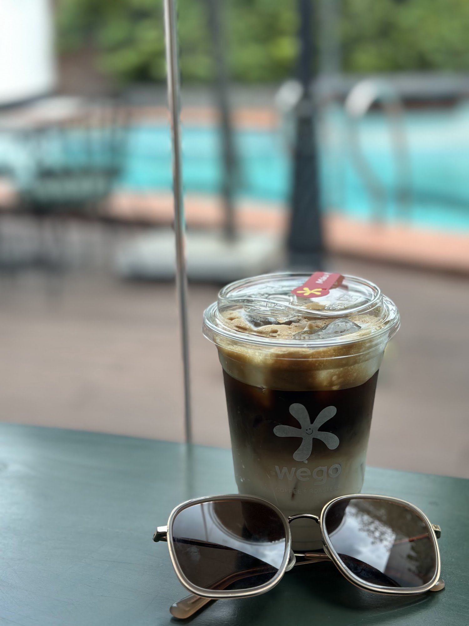 A close-up of a cold coffee drink in a plastic cup from 'We Go Coffee & Tea' with sunglasses resting on the table, set against an out-of-focus backgro