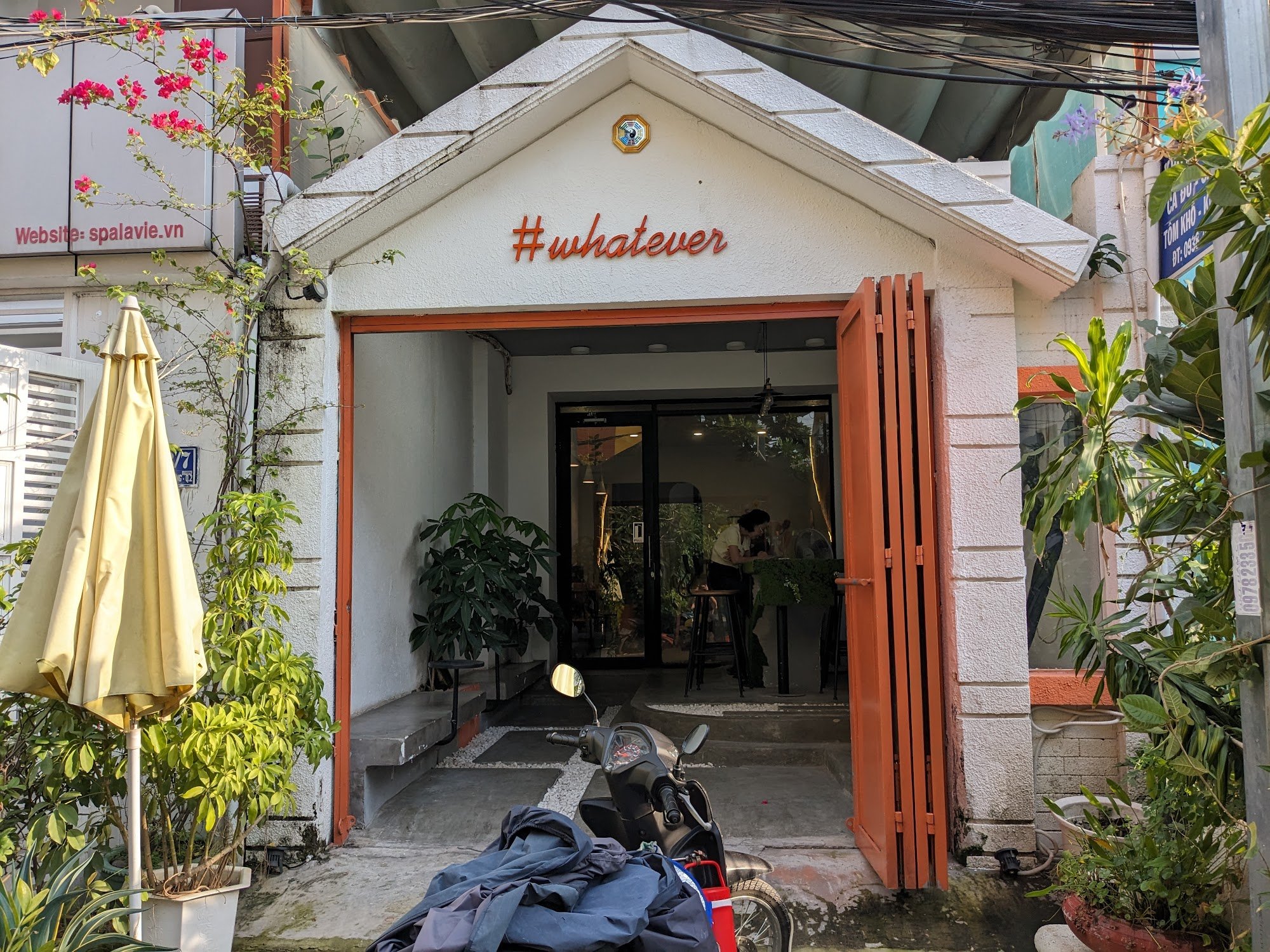 The image shows an exterior view of a cafe named '#whatevers' located in Thao Dien, Saigon. The entrance is marked by a white facade with orange accen
