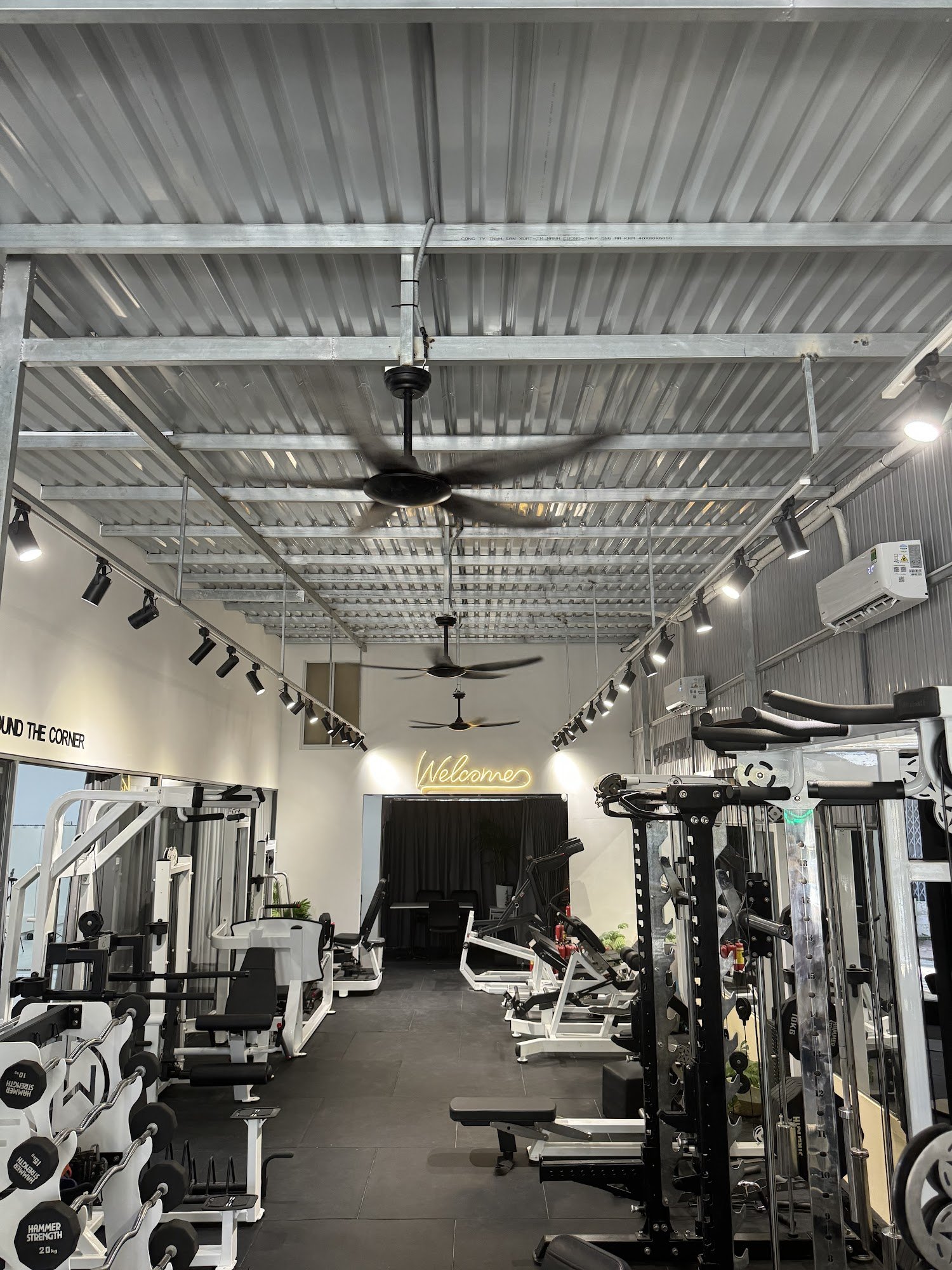 The image shows an interior view of a modern gym located in Thao Dien, Saigon. The space is well-lit with industrial-style lighting fixtures and featu