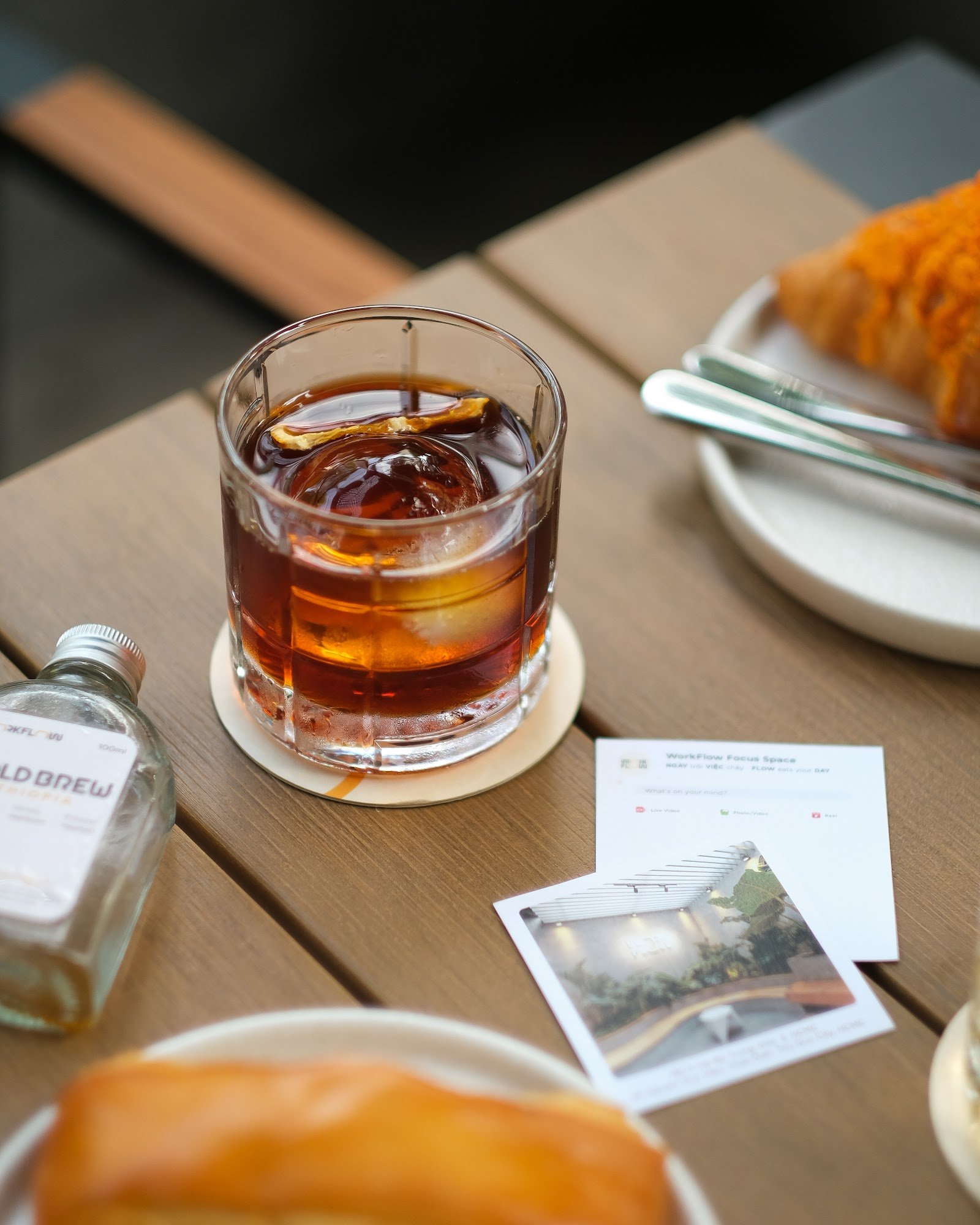 A close-up of a glass containing an amber-colored drink on a wooden table, with various items around it including a bottle labeled 'OLD BREW', a plate