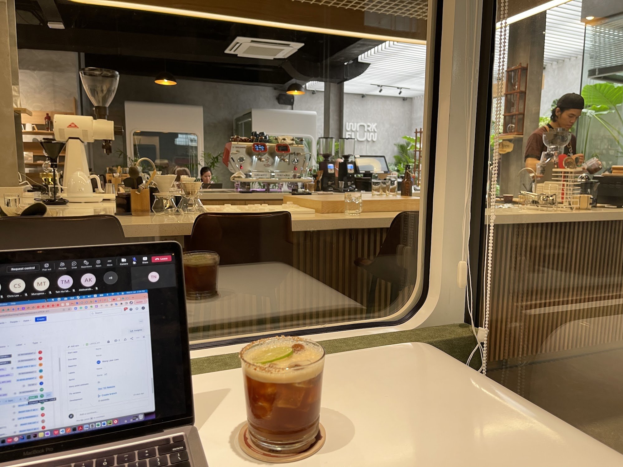 A modern café interior in Thao Dien, Saigon featuring a white table with a laptop displaying a work interface, a glass of iced coffee with lime, a gla