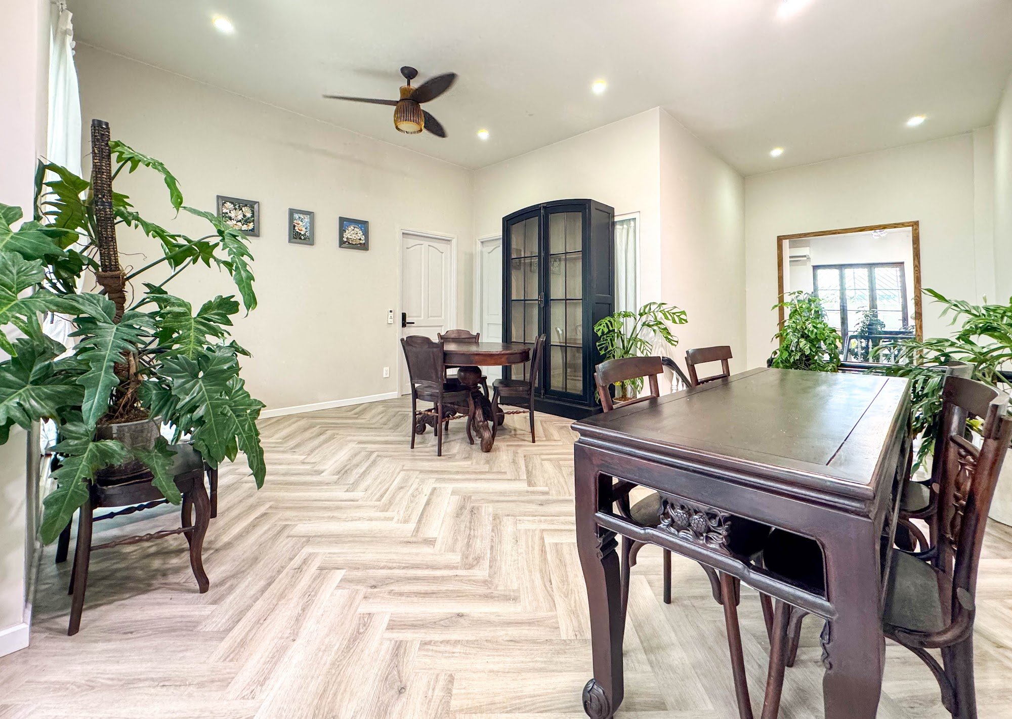A spacious dining area with herringbone-patterned light wood flooring. Features two dark wooden dining tables with matching chairs, a large potted pla
