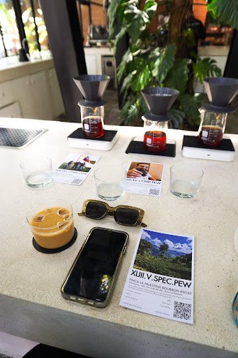 A counter in a café with three pour-over coffee setups (each with a cone and glass), two empty glasses, a glass of coffee with foam, a smartphone, sun