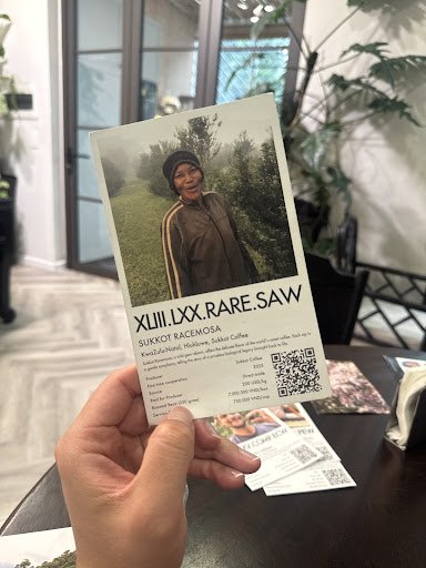 A hand holding a promotional card featuring a photograph of a person in a forested area, with text 'XJIN.IXX.RARE.SAW' and 'SUKKOT KACOMASA', a QR cod