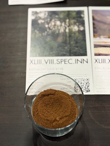 A close-up of a small glass bowl containing brown coffee grounds placed on a dark surface, with a printed menu behind it. The menu features text inclu