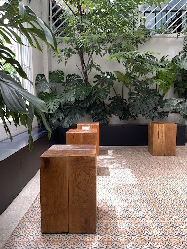 The image depicts an interior space of a venue in Thao Dien, Saigon, featuring several wooden stools arranged on a patterned tile floor. Lush green pl