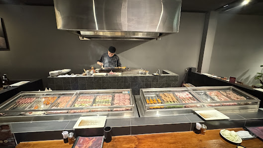 The image shows an interior view of a modern restaurant or food court featuring a chef preparing dishes at the counter with various types of meat and