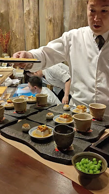 A chef in a white uniform is serving dishes on black trays at an indoor dining venue.