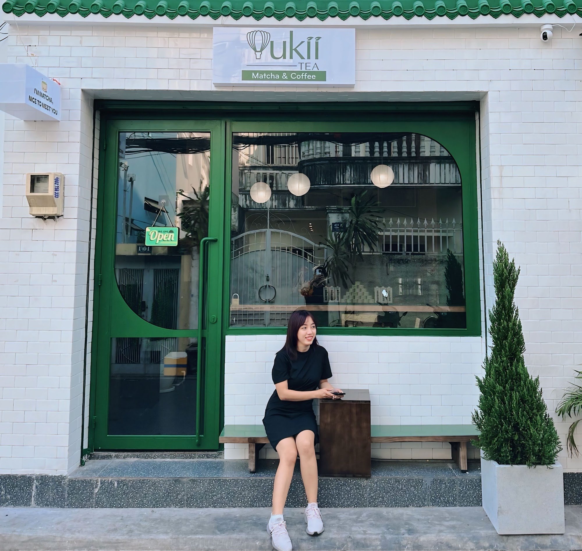 A woman is sitting outside a cafe named 'Pukii Tea' which specializes in matcha and coffee.