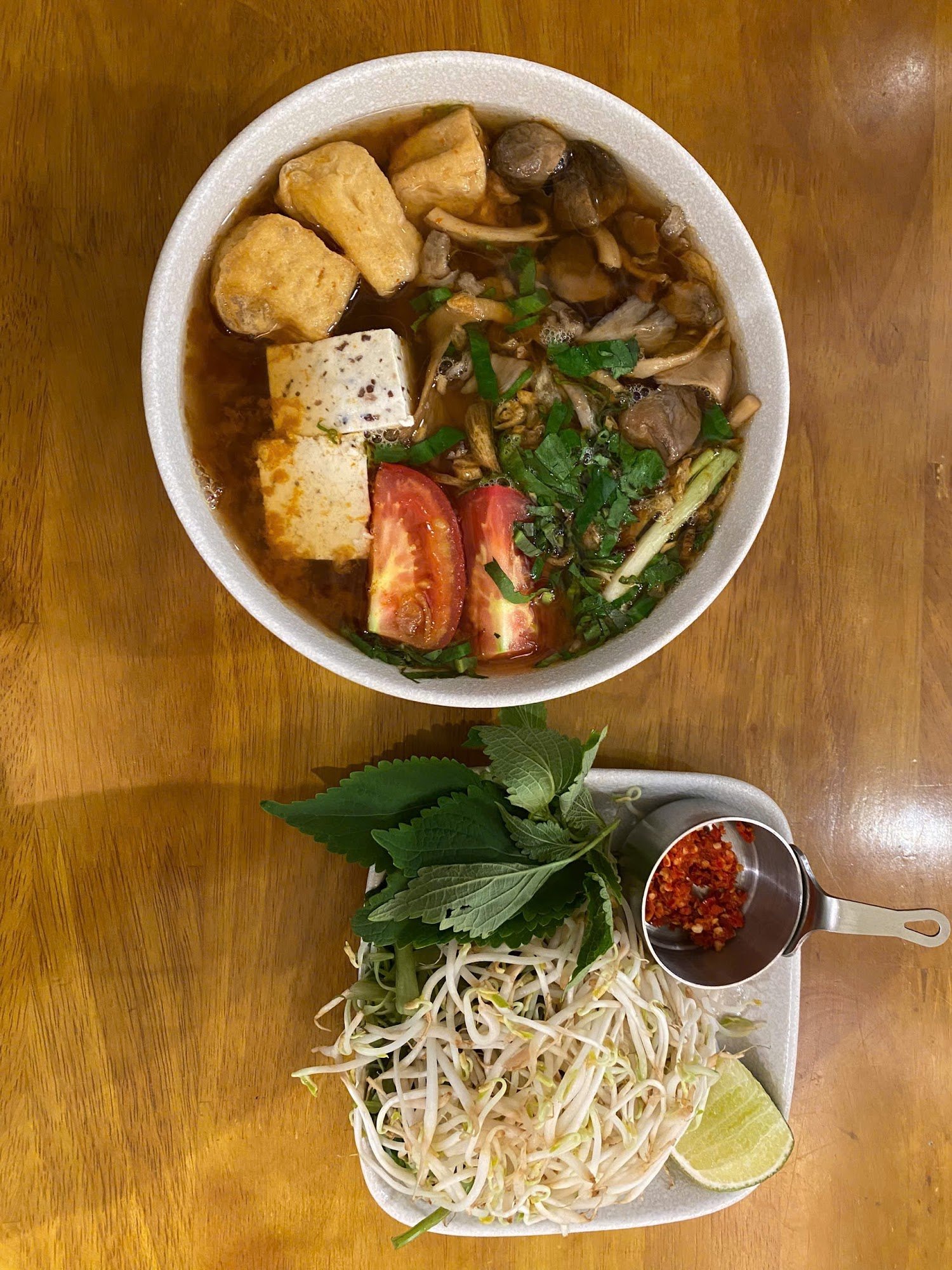 A bowl of soup containing tofu, mushrooms, and leafy greens is accompanied by a side dish consisting of bean sprouts with fresh herbs like mint and ba