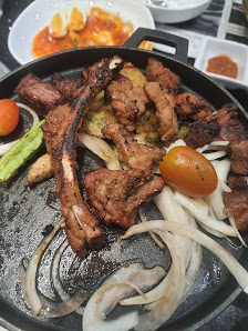 A close-up of a grilled meat dish served on a black pan, featuring what appears to be lamb chops with charred edges and some vegetables like onions an