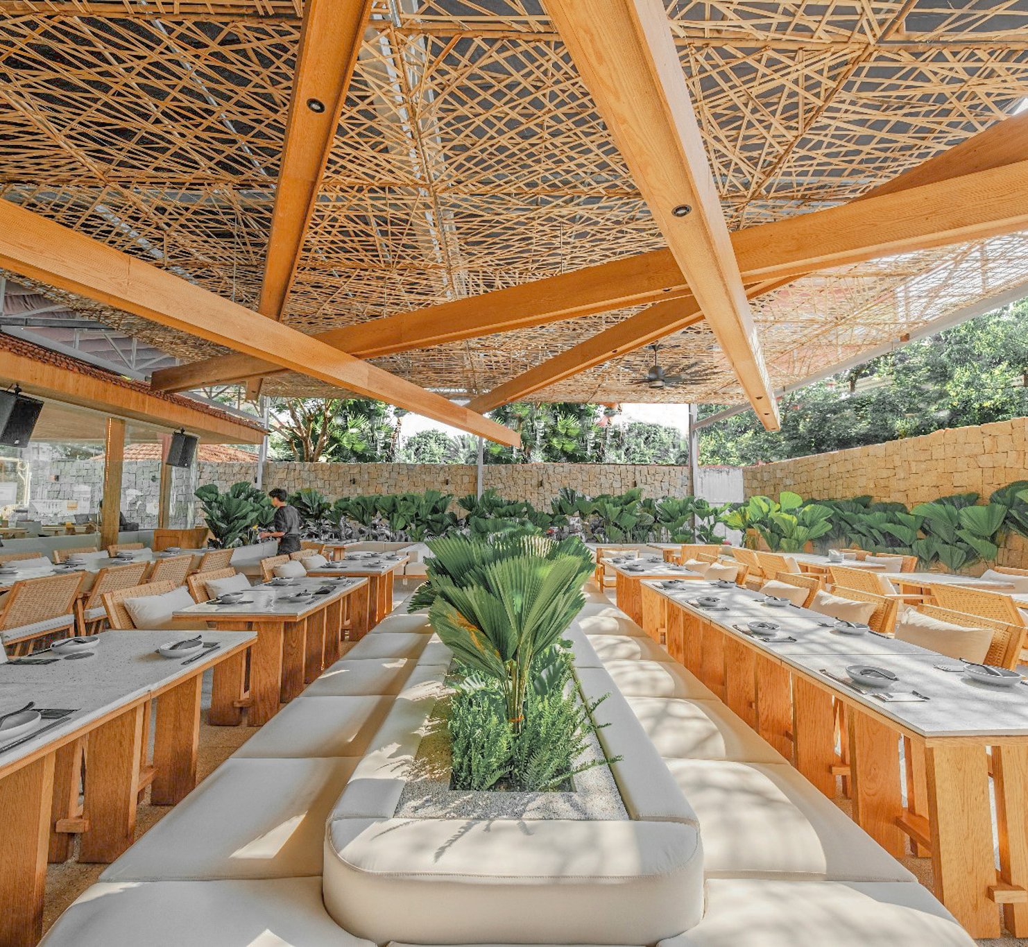 An outdoor dining venue with wooden tables and chairs, a central cushioned seating area, a thatched roof structure, and lush greenery. Stone walls in 