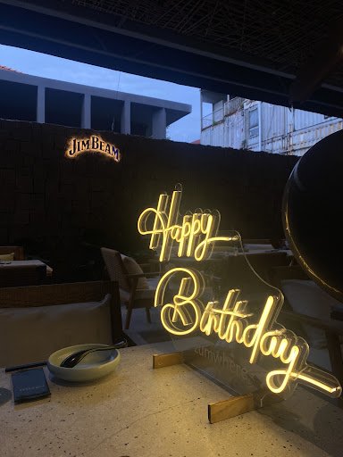 The image depicts an indoor venue in Thao Dien, Saigon, featuring a glowing neon sign that reads 'Happy Birthday' in cursive script. The sign is place