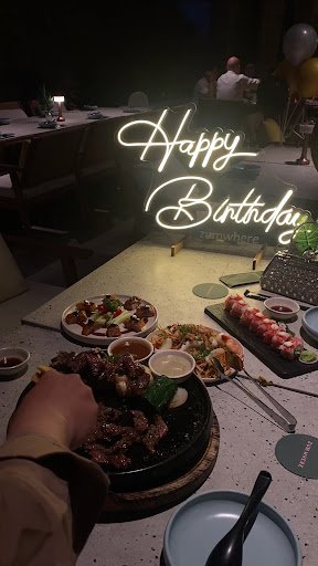 A birthday celebration scene at a restaurant in Thao Dien, Saigon. The image shows a table with various dishes including a sizzling plate of meat (lik