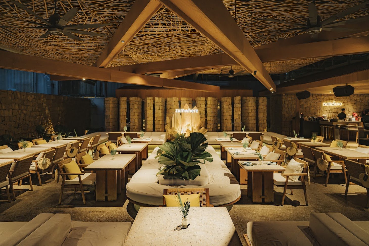 A spacious indoor dining venue with wooden tables, wicker chairs with beige cushions, and a central seating area featuring a large tropical plant. The