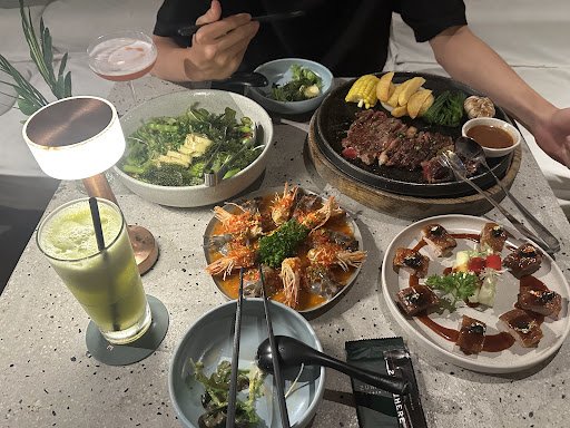 A dining table in a restaurant in Thao Dien, Saigon, set with various dishes including grilled meat on a stone plate, shrimp dish, leafy greens, small
